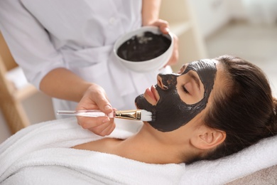 Cosmetologist applying black mask onto woman's face in spa salon