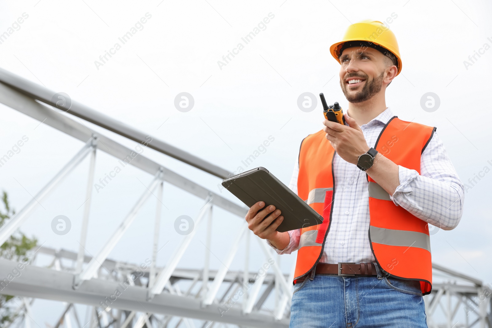 Photo of Professional engineer with tablet and walkie talkie near high voltage tower construction outdoors. Installation of electrical substation