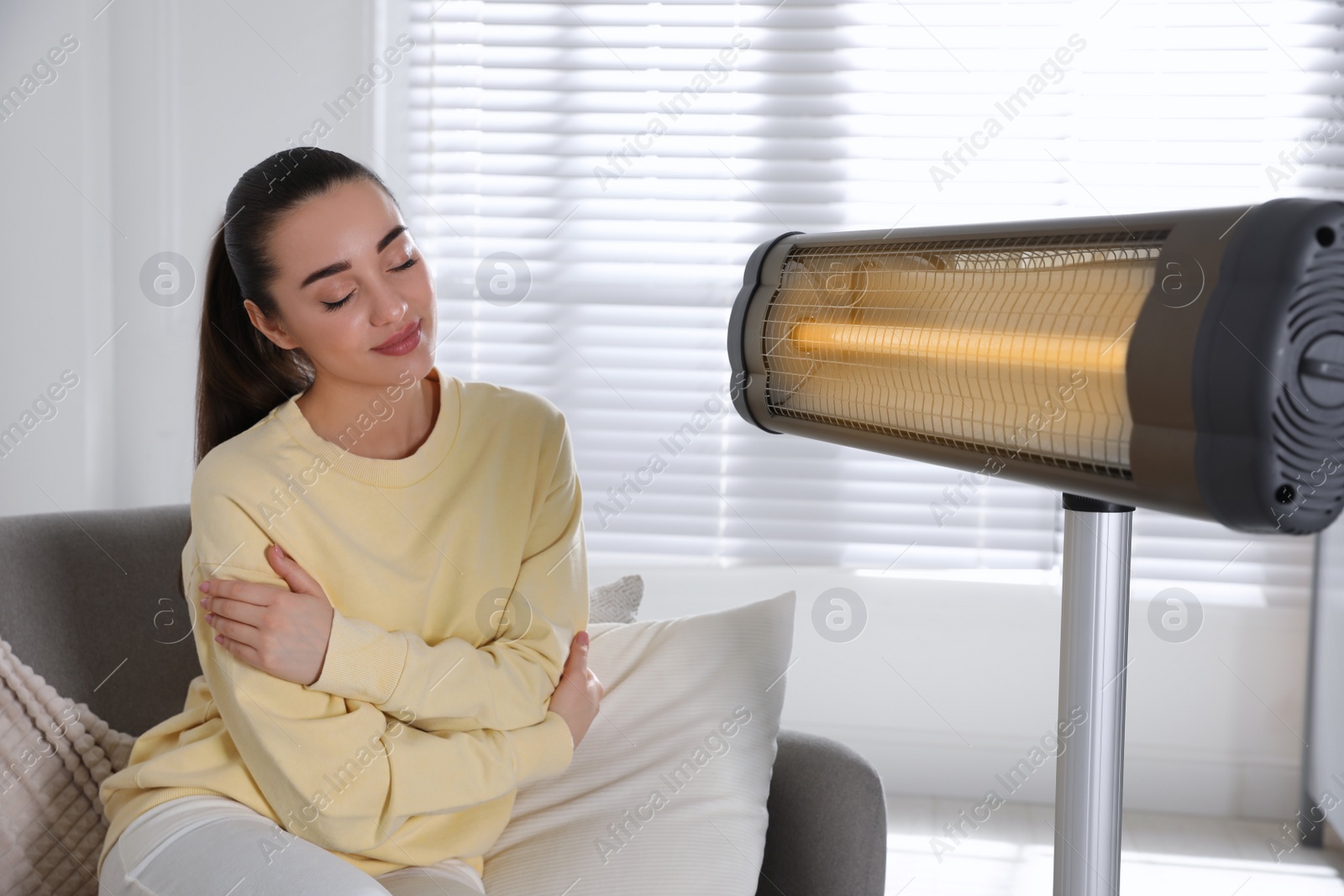 Photo of Young woman sitting on sofa near electric heater at home