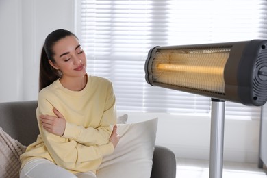 Photo of Young woman sitting on sofa near electric heater at home