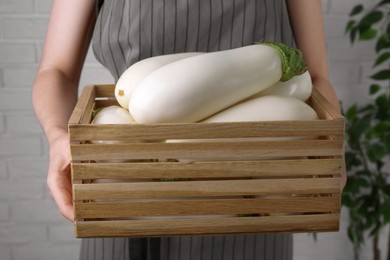 Photo of Woman holding wooden crate with white eggplants near brick wall, closeup