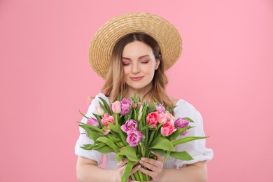 Happy young woman in straw hat holding bouquet of beautiful tulips on pink background