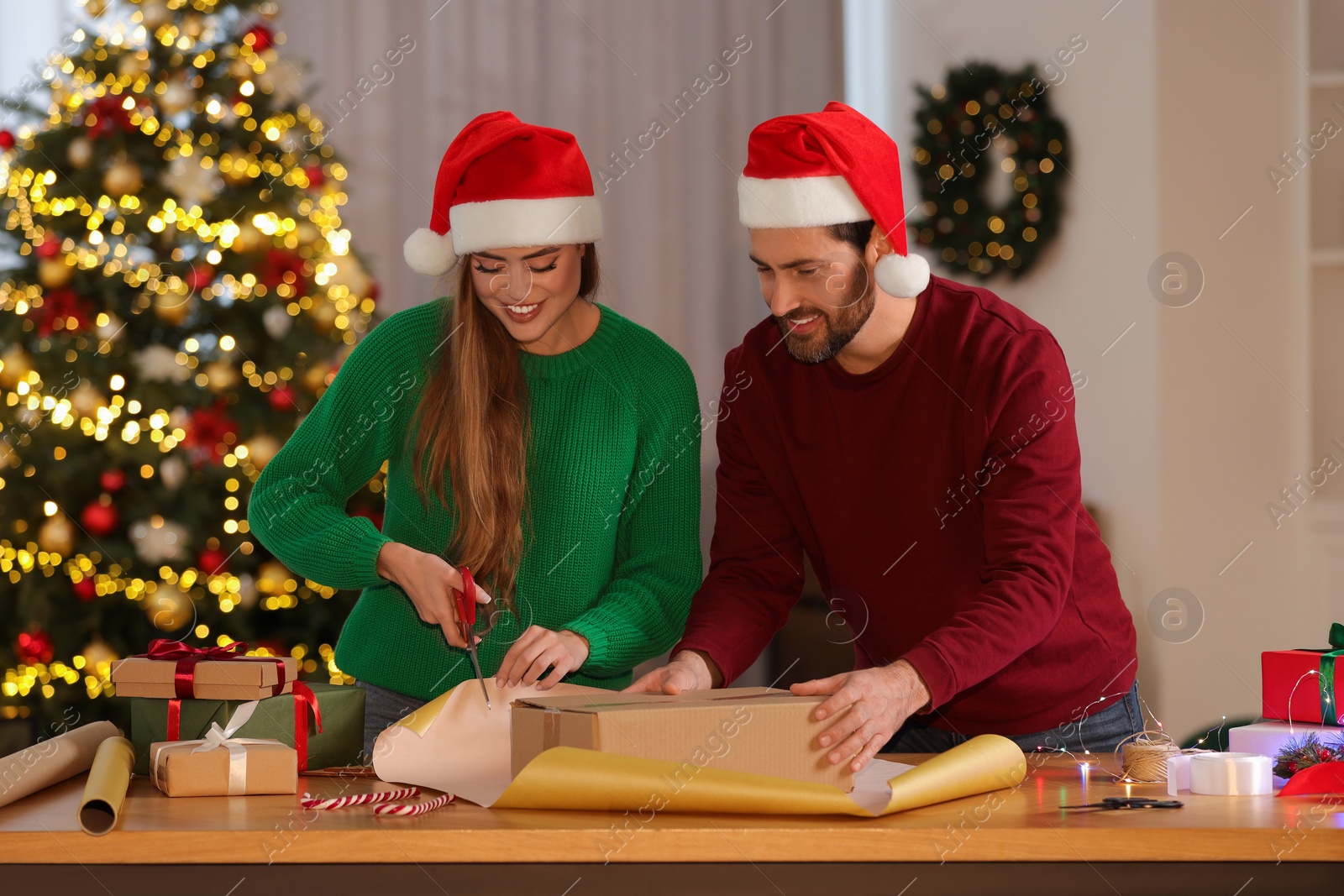 Photo of Happy couple in Santa hats decorating Christmas gift with wrapping paper at table in room