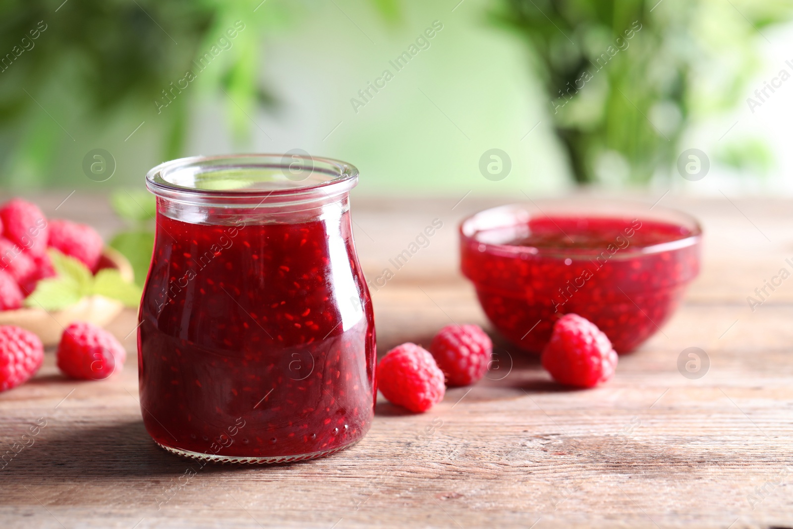 Photo of Glass jar and bowl of sweet jam with ripe raspberries on wooden table against blurred background. Space for text