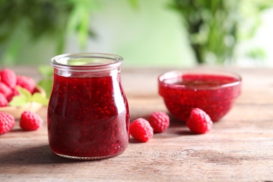 Photo of Glass jar and bowl of sweet jam with ripe raspberries on wooden table against blurred background. Space for text