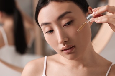 Beautiful young woman applying cosmetic serum onto her face in bathroom, closeup