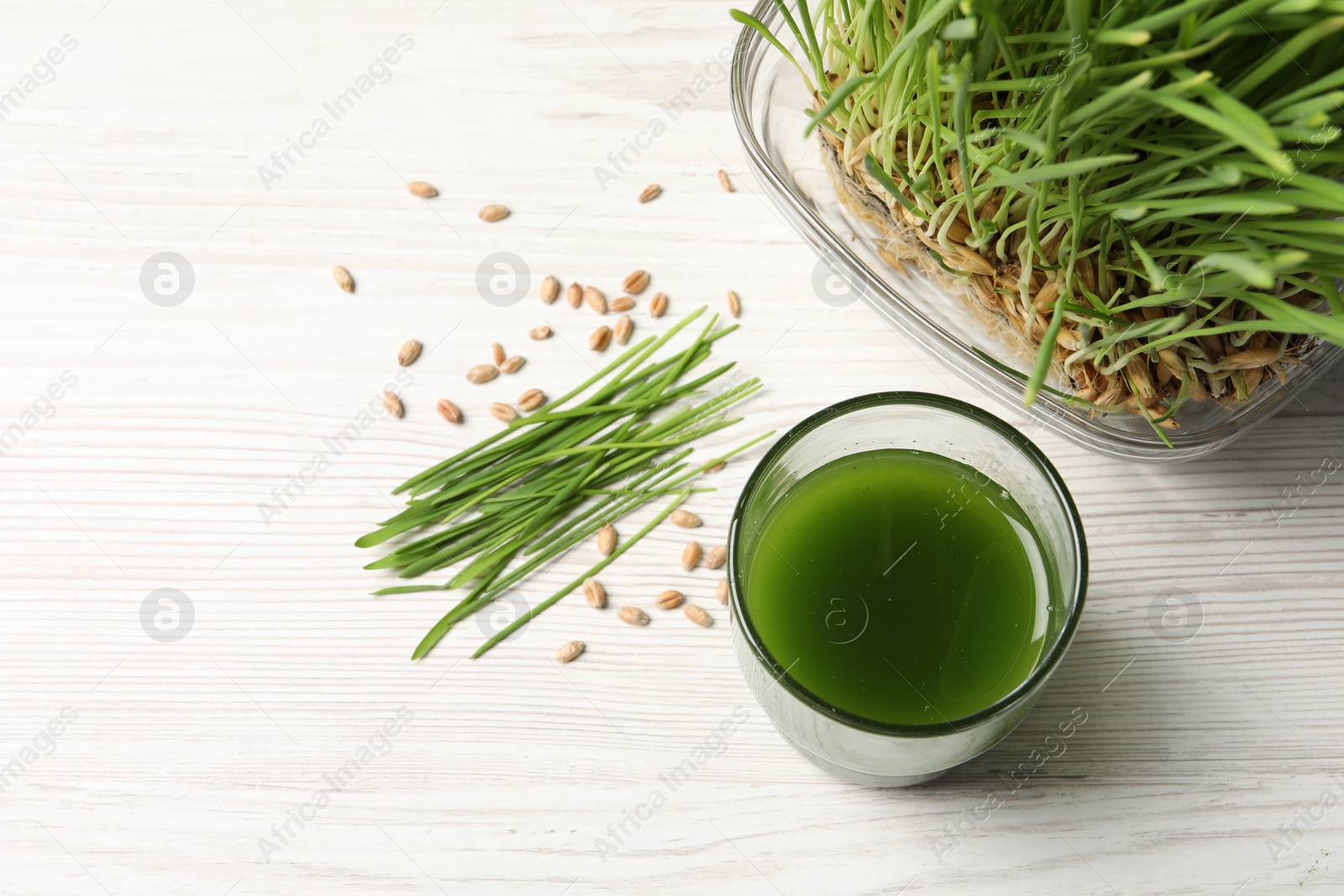 Photo of Wheat grass drink in glass, seeds and fresh green sprouts on white wooden table, flat lay