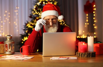 Photo of Santa Claus using laptop at his workplace in room with Christmas tree