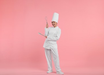 Photo of Happy professional confectioner in uniform holding whisk and spatula on pink background