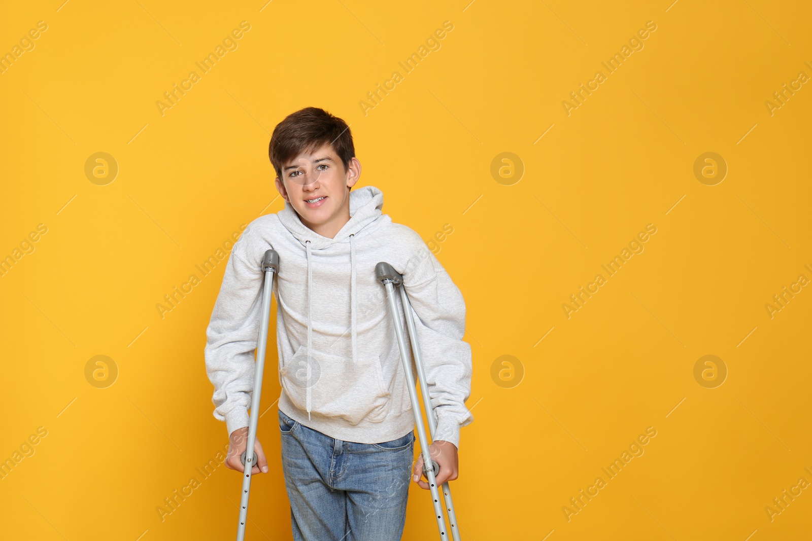 Photo of Teenage boy with injured leg using crutches on yellow background, space for text