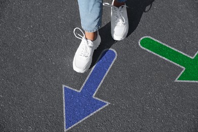 Image of Choice of way. Woman walking to drawn mark on road, closeup. Green and blue arrows pointing in different directions