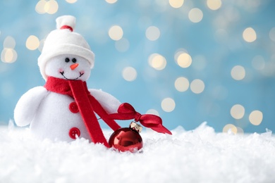 Photo of Snowman toy and Christmas ball on snow against blurred festive lights, closeup. Space for text