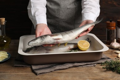 Woman putting raw sea bass fish into baking tray at wooden table, closeup