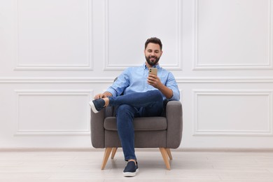 Photo of Handsome man with smartphone sitting in armchair near white wall indoors