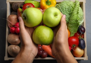 Farmer with wooden crate full of different vegetables and fruits at grey table, top view. Harvesting time