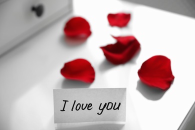 Paper with text I Love You and rose petals on white table