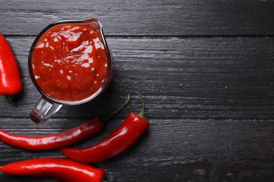 Photo of Jug of chili sauce and pepper on wooden table, top view. Space for text
