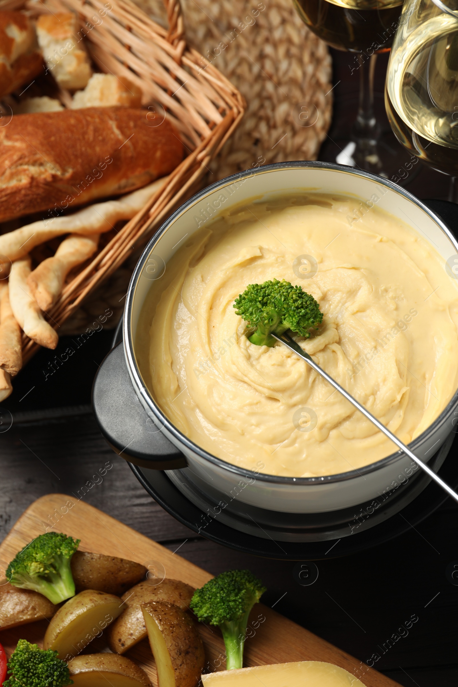 Photo of Dipping piece of broccoli into fondue pot with melted cheese at table with snacks, flat lay