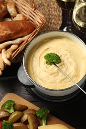 Photo of Dipping piece of broccoli into fondue pot with melted cheese at table with snacks, flat lay