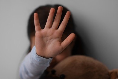 Photo of Child abuse. Little girl with teddy bear doing stop gesture on light grey background, selective focus