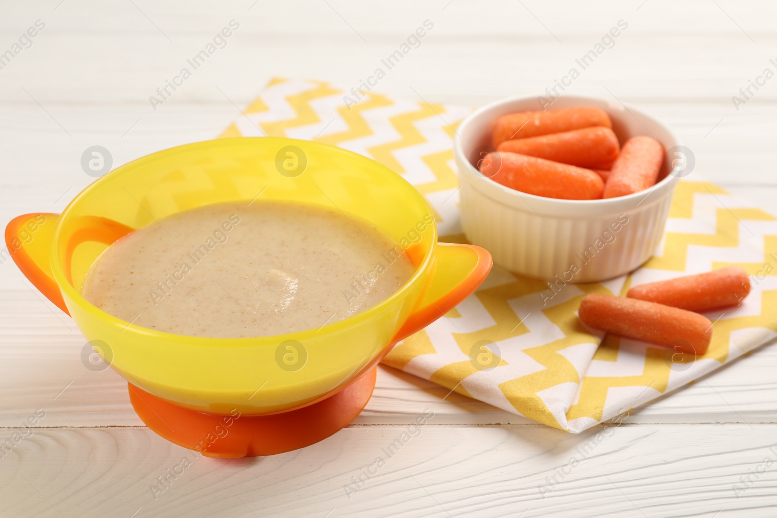 Photo of Baby food. Puree in bowl and small carrots on white wooden table