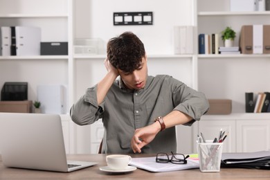 Photo of Emotional young man checking time while working at table in office. Deadline concept