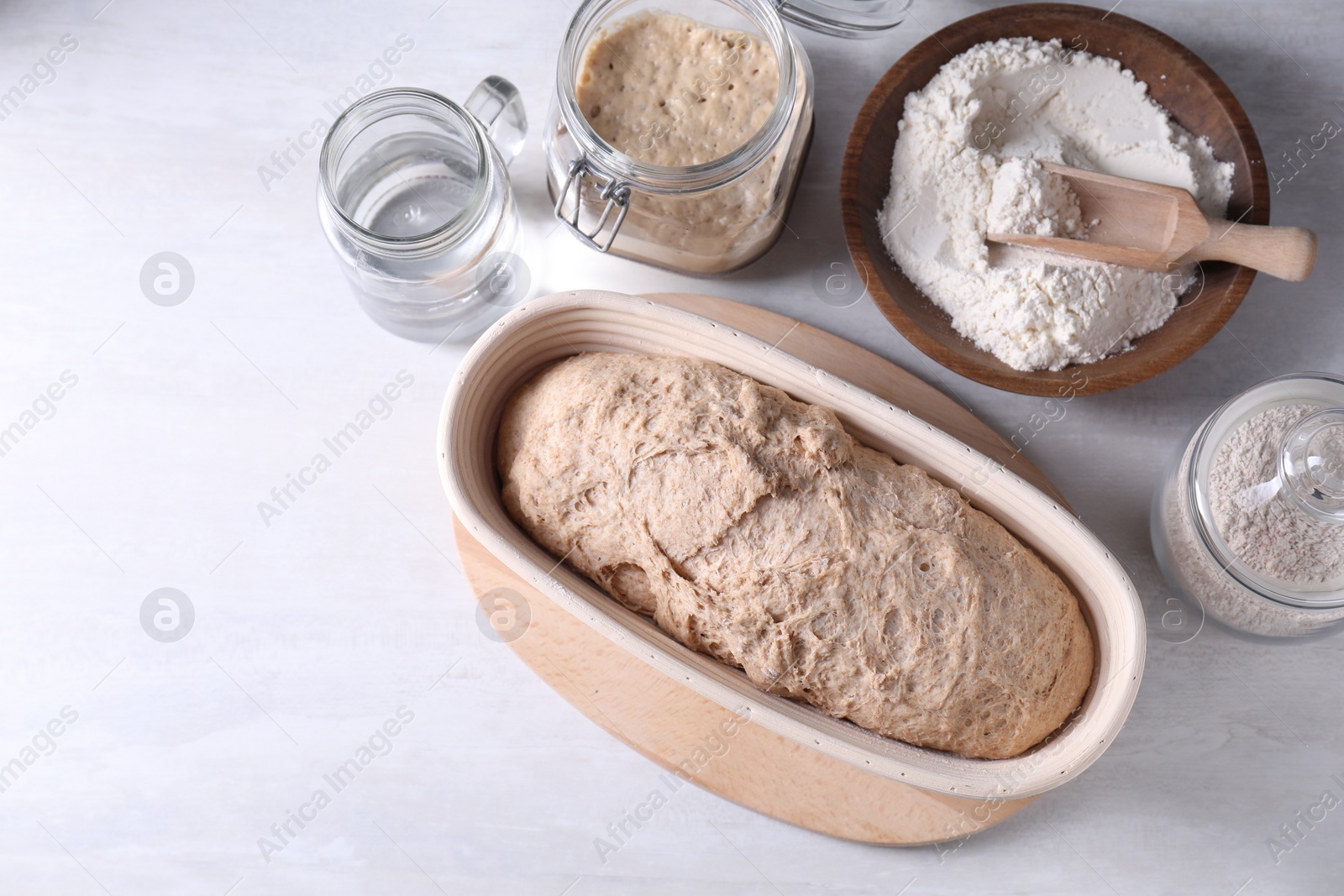 Photo of Fresh sourdough in proofing basket, flour and water on light table, flat lay. Space for text