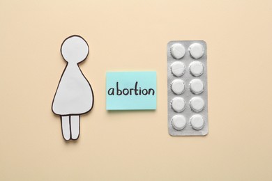 Photo of Note with word Abortion, pills and pregnant woman paper cutout on beige background, flat lay