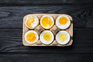 Photo of Boiled chicken eggs of different readiness stages in carton on black wooden table, top view
