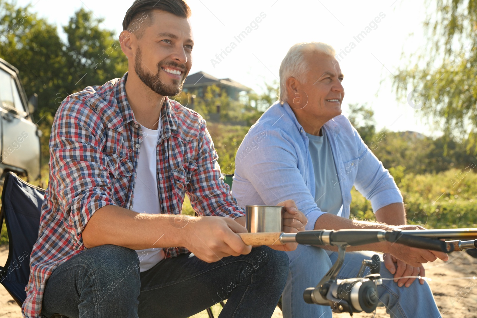 Photo of Father and adult son fishing together from riverside on sunny day