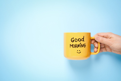 Photo of Woman holding cup of coffee with words GOOD MORNING on light blue background, closeup. Space for text