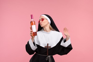 Happy woman in nun habit and sunglasses holding bottle of wine on pink background
