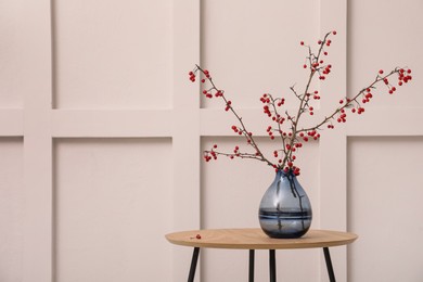 Photo of Hawthorn branches with red berries in vase on wooden table near white wall indoors, space for text. Interior element