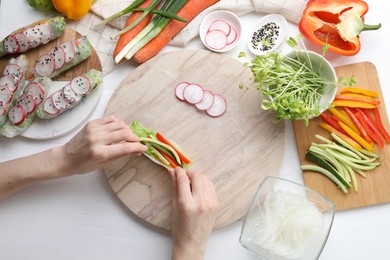 Photo of Woman wrapping spring roll at white wooden table with products, top view