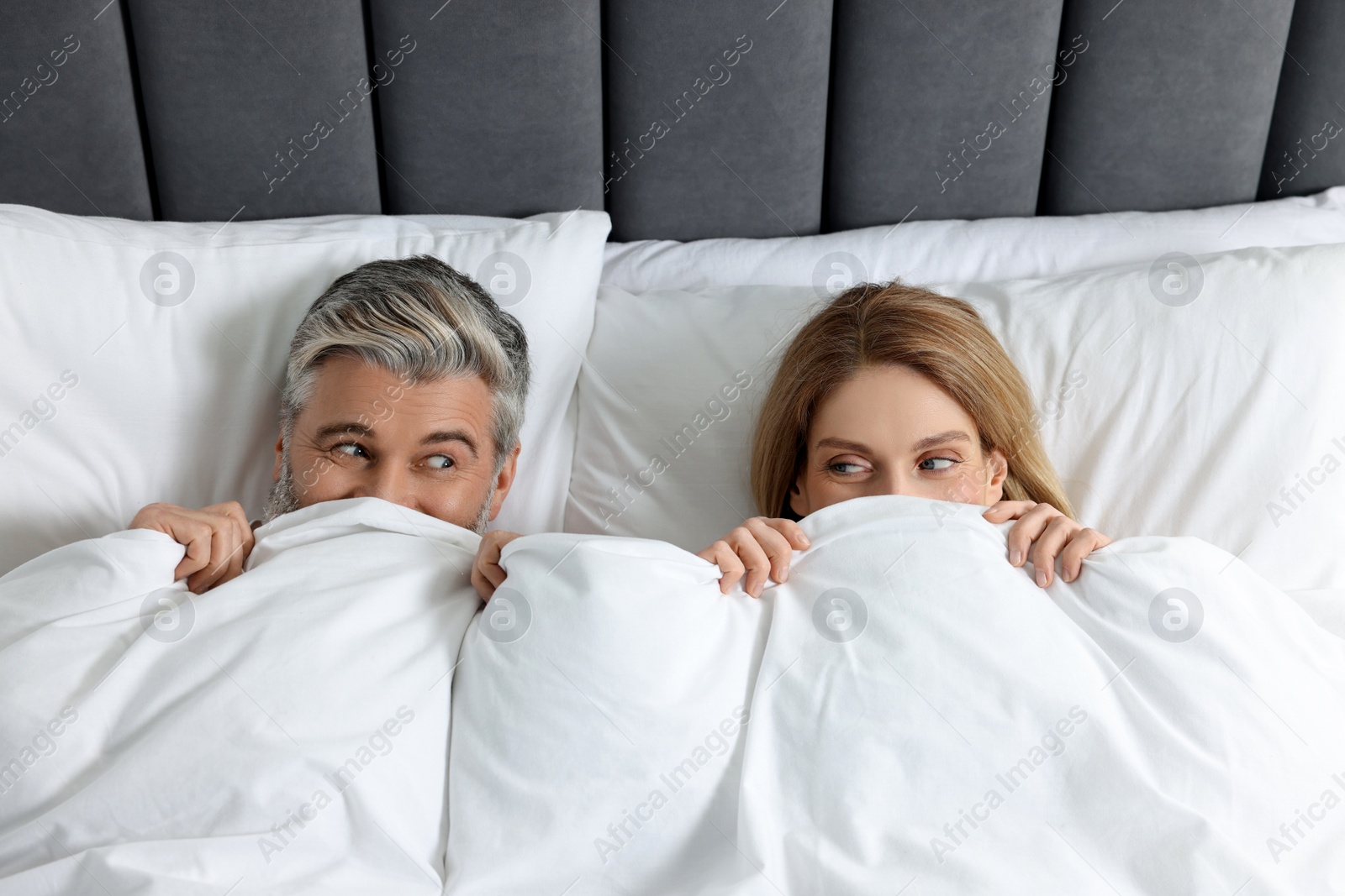 Photo of Lovely mature couple hiding together under blanket in bed at home, above view