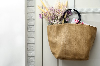 Stylish beach bag with beautiful bouquet of wildflowers and magazine hanging on white wooden door. Space for text