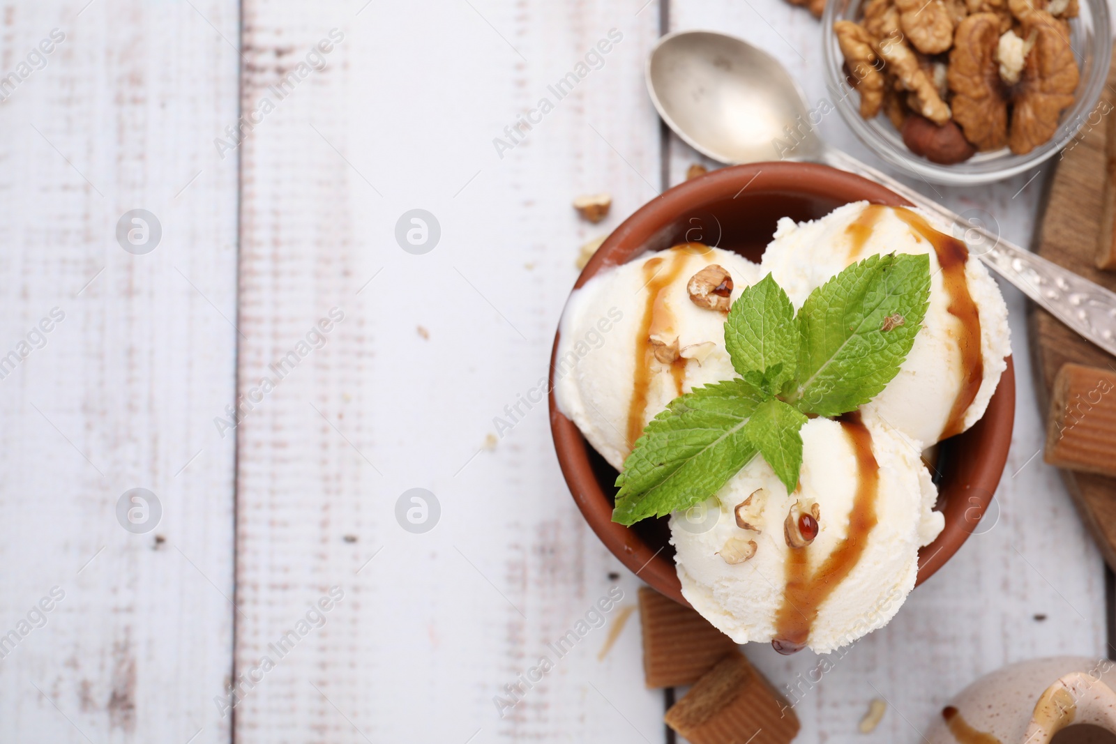 Photo of Bowl of tasty ice cream with caramel sauce, mint and nuts on white wooden table, flat lay. Space for text