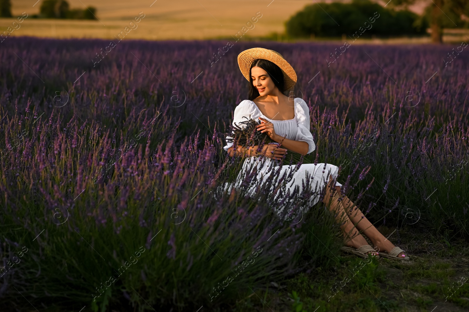 Photo of Beautiful young woman with bouquet sitting in lavender field at sunset