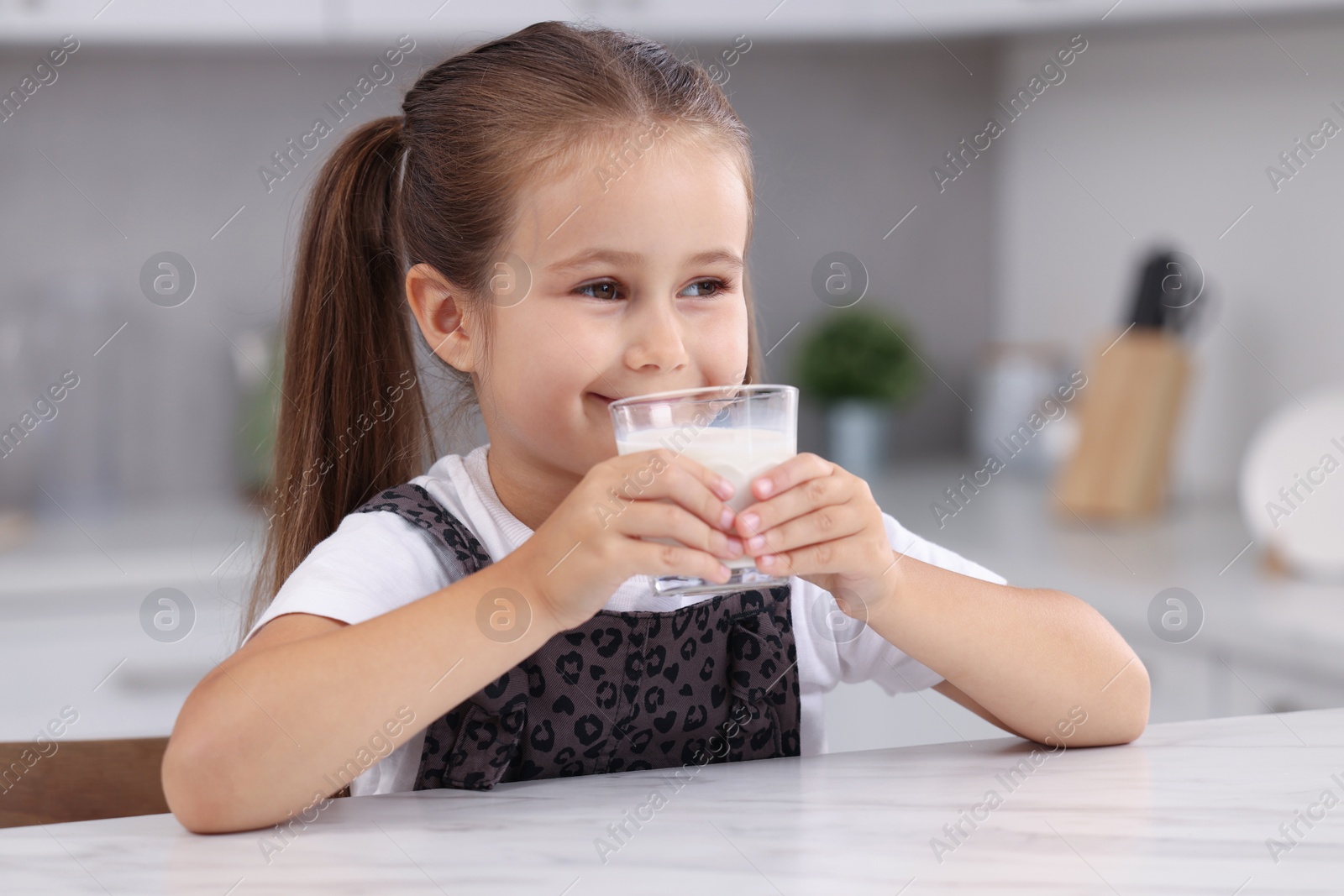 Photo of Cute girl drinking fresh milk from glass at white table in kitchen