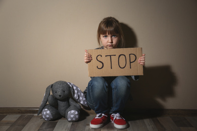 Abused little girl with sign STOP near beige wall. Domestic violence concept