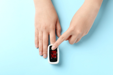Woman using fingertip pulse oximeter on light blue background, top view