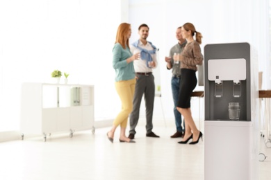 Photo of Modern water cooler with glass and blurred office employees on background. Space for text