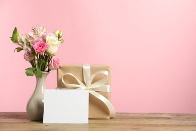 Happy Mother's Day. Gift box, blank card and bouquet of beautiful flowers in vase on wooden table against pink background. Space for text