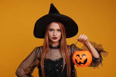Photo of Young woman in scary witch costume with pumpkin bucket on orange background. Halloween celebration
