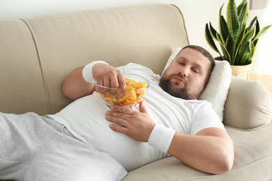 Photo of Lazy overweight man eating chips on sofa at home