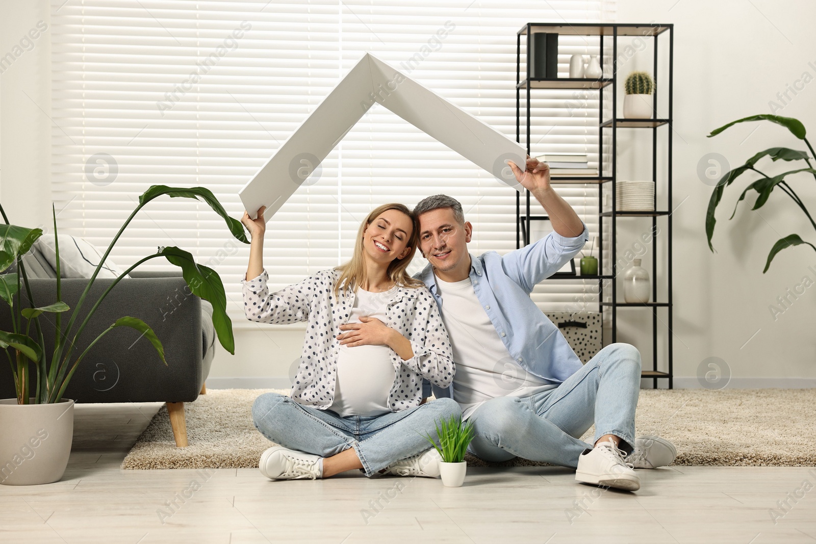 Photo of Young family housing concept. Pregnant woman with her husband sitting under cardboard roof on floor at home