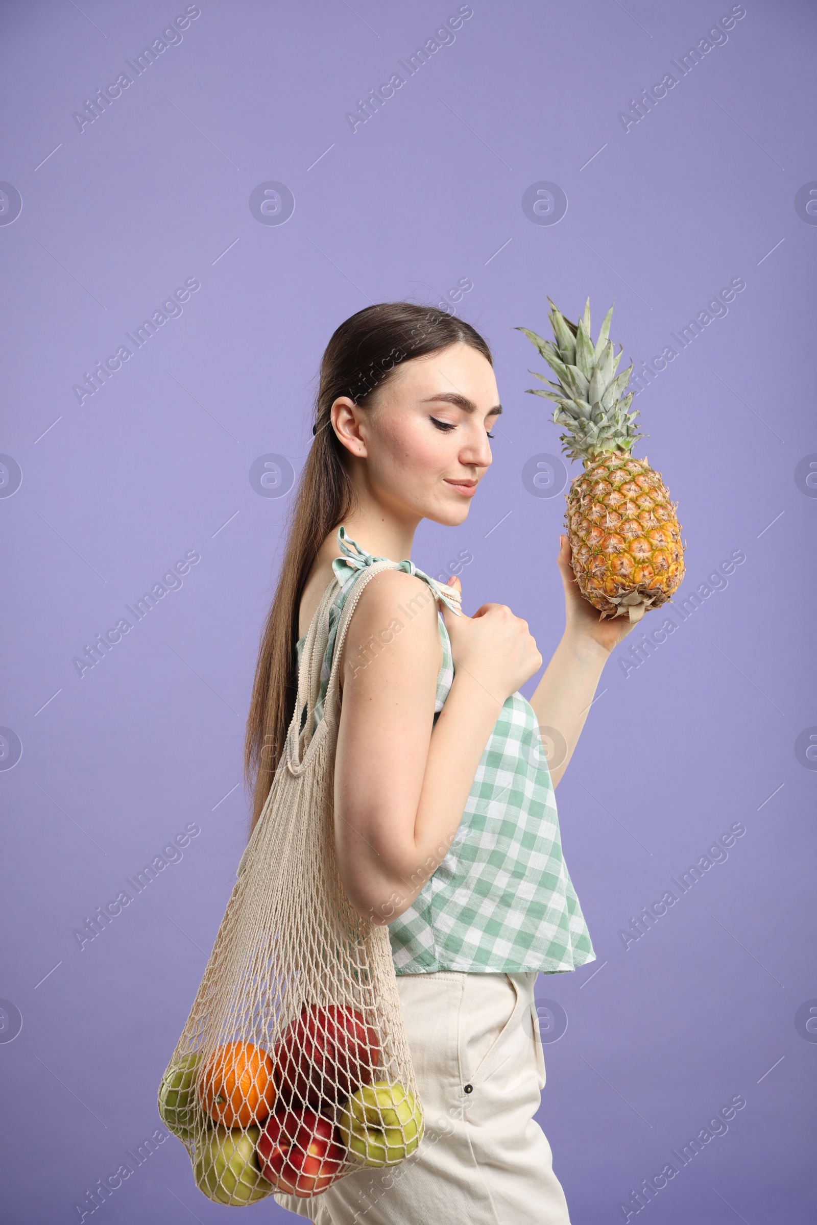 Photo of Woman with string bag of fresh fruits holding pineapple on violet background