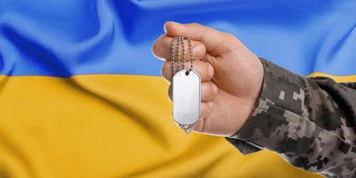 Image of Closeup view of soldier in uniform holding military ID tag and Ukrainian flag on background. Banner design