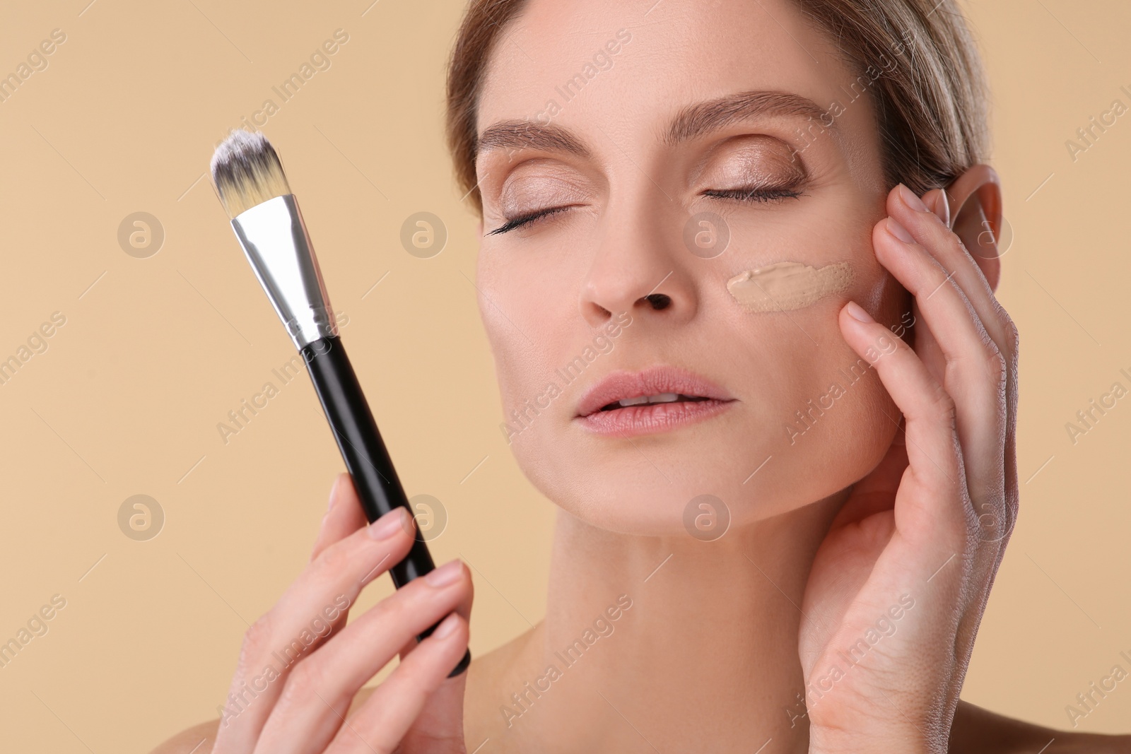 Photo of Woman with swatch of foundation holding makeup brush on beige background