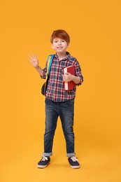 Photo of Happy schoolboy with backpack and book waving hello on orange background